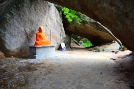 A person dressed in bright orange robes sits cross-legged in meditation on a stone bench against a large rock wall. The scene is set outdoors, beneath an arch formed by a massive boulder, with greenery visible in the background. There are some signs nearby on the ground.
