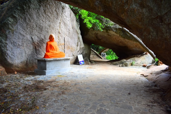 A person dressed in bright orange robes sits cross-legged in meditation on a stone bench against a large rock wall. The scene is set outdoors, beneath an arch formed by a massive boulder, with greenery visible in the background. There are some signs nearby on the ground.