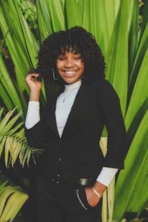 A person with curly hair is smiling while standing in front of large green leaves. The person is wearing a black blazer over a white garment, with one hand in their pocket and the other touching their hair.