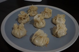 Eight steamed dumplings with a delicate, pleated design are arranged in a circle on a round, blue-gray plate. The dumplings have a slightly shiny appearance, suggesting they're freshly cooked, and they sit on a dark surface.