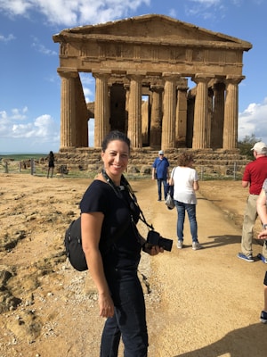 A woman with a camera around her neck stands smiling in front of an ancient, weathered temple with strong vertical columns. The ground is dusty, and the sky is partly cloudy. Other tourists can be seen around the temple, some closer to the structure itself.