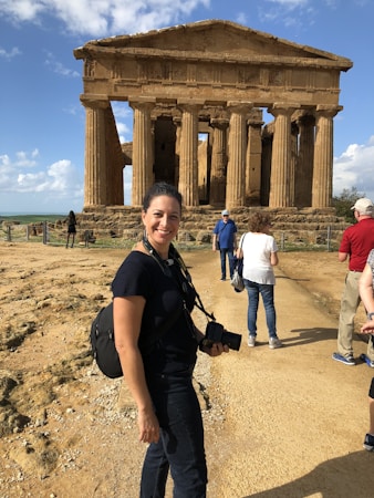 A woman with a camera around her neck stands smiling in front of an ancient, weathered temple with strong vertical columns. The ground is dusty, and the sky is partly cloudy. Other tourists can be seen around the temple, some closer to the structure itself.