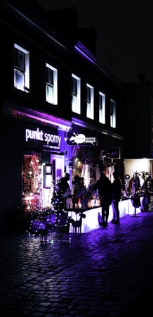 A night scene featuring an illuminated storefront with the sign 'punkt sporny' in bright lights. The windows above the shop glow with a soft violet hue. A group of people stands near tables that are lit from underneath, creating a striking contrast against the dark, cobblestone street. Decorative lights are strung around the entrance, adding a festive touch.