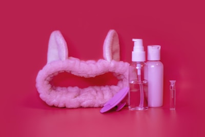 A collection of cosmetic items including a fluffy pink headband with bunny ears, a clear spray bottle, a small squeeze bottle, a translucent vial, and a pink facial cleansing brush arranged against a vibrant pink background.