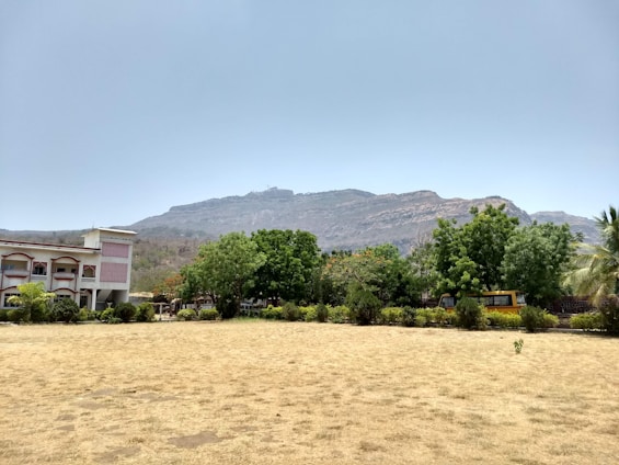 A suburban landscape featuring a large grassy field and a two-story building with a red and white facade on the left. The scene includes several mature trees and a visible mountain range in the background under a clear blue sky. A yellow vehicle is parked among the trees.