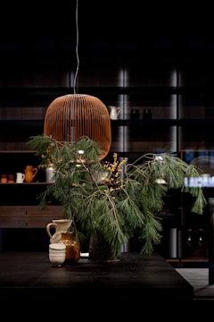 A sophisticated interior setting featuring a modern, wire-framed pendant light hanging above a dark table. The table is adorned with a lush arrangement of pine branches and yellow berries in a vase, accompanied by several ceramic mugs and pitchers. The background consists of shelves displaying various kitchenware in a dimly lit room.