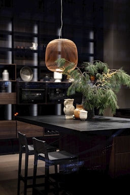 A modern kitchen features a dark-toned island with two chairs. On the island, there are ceramic jugs and vases, along with a green pine branch centerpiece. Above the island hangs a wooden pendant light with a slatted design. The background has open shelves and integrated kitchen appliances.