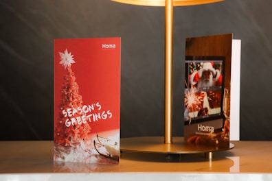 Two holiday greeting cards are displayed on a beige surface under a gold lamp. The card on the left has a red background featuring a red Christmas tree topped with a white star and the words 'Season's Greetings' in white. The card on the right shows a festive scene with a Santa Claus image, fireworks, and a champagne glass.