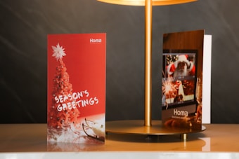 Two holiday greeting cards are displayed on a beige surface under a gold lamp. The card on the left has a red background featuring a red Christmas tree topped with a white star and the words 'Season's Greetings' in white. The card on the right shows a festive scene with a Santa Claus image, fireworks, and a champagne glass.