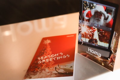 Two greeting cards are displayed on a table. One card is red with a Christmas tree and the words 'Season's Greetings'. The other card shows a digital screen with a Santa figure and a sparkler next to a glass of champagne.