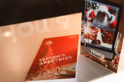 Two greeting cards are displayed on a table. One card is red with a Christmas tree and the words 'Season's Greetings'. The other card shows a digital screen with a Santa figure and a sparkler next to a glass of champagne.