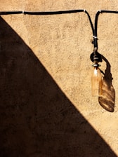 A textured beige wall with a prominent diagonal shadow. An old, weathered glass insulator is attached to the wall, casting its own shadow. Black wires run horizontally and vertically, creating a pattern against the wall.