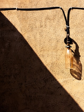 A textured beige wall with a prominent diagonal shadow. An old, weathered glass insulator is attached to the wall, casting its own shadow. Black wires run horizontally and vertically, creating a pattern against the wall.