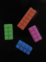 Four colorful toy building blocks are placed on a black background. The blocks are rectangular with rounded pegs on top. They are in different colors: orange, pink, blue, and green.