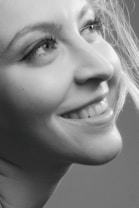 A close-up portrait of a person with long eyelashes and a smile, shown in black and white. The image captures the side profile with light highlighting the skin and hair details.