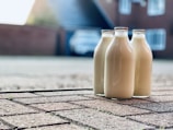 Three glass bottles filled with milk are placed on a paved driveway. In the background, a house and a blurry car can be seen, indicating a residential setting. The perspective is close to the ground, focusing on the bottles, with the background softly out of focus.