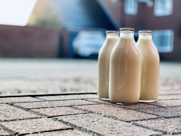 Three glass bottles filled with milk are placed on a paved driveway. In the background, a house and a blurry car can be seen, indicating a residential setting. The perspective is close to the ground, focusing on the bottles, with the background softly out of focus.