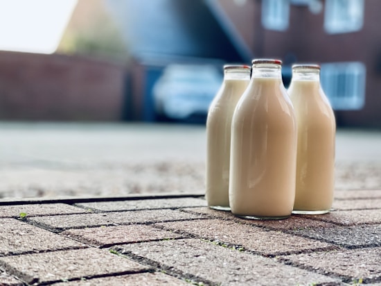 Three glass bottles filled with milk are placed on a paved driveway. In the background, a house and a blurry car can be seen, indicating a residential setting. The perspective is close to the ground, focusing on the bottles, with the background softly out of focus.