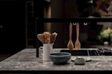 A modern kitchen countertop made of stone with a white ceramic holder containing wooden cooking utensils. In the foreground, there is a blue bowl and a small green cup. Three wooden kitchen utensils are hanging on a black metal bar.