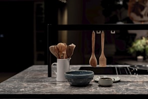 A modern kitchen countertop made of stone with a white ceramic holder containing wooden cooking utensils. In the foreground, there is a blue bowl and a small green cup. Three wooden kitchen utensils are hanging on a black metal bar.