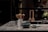 A modern kitchen countertop made of stone with a white ceramic holder containing wooden cooking utensils. In the foreground, there is a blue bowl and a small green cup. Three wooden kitchen utensils are hanging on a black metal bar.
