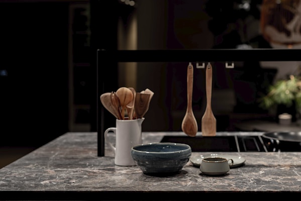 A modern kitchen countertop made of stone with a white ceramic holder containing wooden cooking utensils. In the foreground, there is a blue bowl and a small green cup. Three wooden kitchen utensils are hanging on a black metal bar.