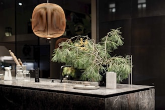 A modern kitchen countertop made of dark marble is adorned with a black vase holding green pine branches and small yellow flowers. A wooden pendant lamp hangs above, casting a warm glow. The countertop also features kitchen items like a white carafe, glass jars, and a striped ceramic cup.