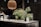 A modern kitchen countertop made of dark marble is adorned with a black vase holding green pine branches and small yellow flowers. A wooden pendant lamp hangs above, casting a warm glow. The countertop also features kitchen items like a white carafe, glass jars, and a striped ceramic cup.
