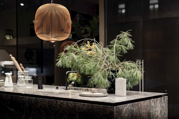 A modern kitchen countertop made of dark marble is adorned with a black vase holding green pine branches and small yellow flowers. A wooden pendant lamp hangs above, casting a warm glow. The countertop also features kitchen items like a white carafe, glass jars, and a striped ceramic cup.