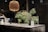 A modern kitchen countertop made of dark marble is adorned with a black vase holding green pine branches and small yellow flowers. A wooden pendant lamp hangs above, casting a warm glow. The countertop also features kitchen items like a white carafe, glass jars, and a striped ceramic cup.