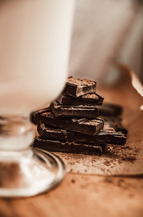 A stack of dark chocolate pieces is placed on a wooden surface. The chocolate is dusted with a fine layer of cocoa powder, and the texture is visible. In the foreground, part of a glass is visible, slightly out of focus, suggesting a rustic, cozy setting.