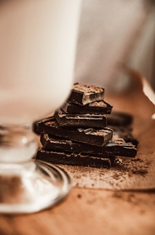 A stack of dark chocolate pieces is placed on a wooden surface. The chocolate is dusted with a fine layer of cocoa powder, and the texture is visible. In the foreground, part of a glass is visible, slightly out of focus, suggesting a rustic, cozy setting.
