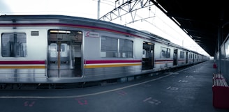 A silver commuter train with red and yellow stripes is stationary at an empty platform. The scene is industrial, with overhead cables and a roof covering the platform. The train doors are open, revealing a well-lit interior. The platform is marked with signage and symbols, and a few red benches are visible.