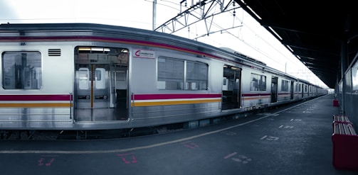 A silver commuter train with red and yellow stripes is stationary at an empty platform. The scene is industrial, with overhead cables and a roof covering the platform. The train doors are open, revealing a well-lit interior. The platform is marked with signage and symbols, and a few red benches are visible.