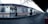 A silver commuter train with red and yellow stripes is stationary at an empty platform. The scene is industrial, with overhead cables and a roof covering the platform. The train doors are open, revealing a well-lit interior. The platform is marked with signage and symbols, and a few red benches are visible.