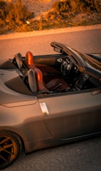 A sports convertible car with its top down, featuring a sleek design and two red leather seats. The steering wheel and dashboard are visible, showcasing modern controls. The car is positioned on a paved road, and the scene is lit by warm, golden sunlight, suggesting a sunset or sunrise. Surrounding the vehicle, dry grass and sparse vegetation add a natural touch to the otherwise urban setting.