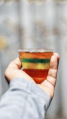 Hand holding a transparent cup filled with golden-yellow passion fruit gelatina, sunlight highlighting its texture.