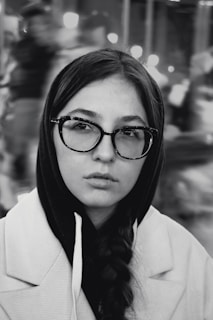 A young person with long braided hair is wearing a hooded sweater under a light-colored coat, and large framed glasses. The background is blurred, suggesting a bustling or busy urban setting at night.