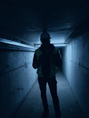 Worker using handheld Synexar gas detector while navigating a narrow pipeline corridor