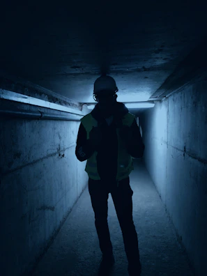 Worker using handheld Synexar gas detector while navigating a narrow pipeline corridor