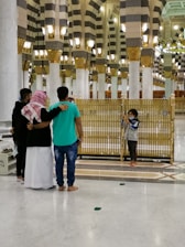 A group of people stands inside a grand mosque with intricate architecture, featuring ornate columns and arches. The floor is polished and people are barefoot. A child is seen by the railing, possibly preparing to take a photo.