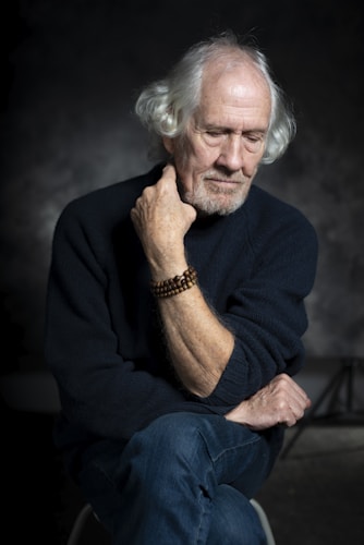 An elderly man with white hair and a thoughtful expression sits against a dark background. He rests his head on one hand, with the other arm crossed, wearing a navy sweater and blue jeans. A wooden bead bracelet adorns his wrist.