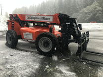 Large orange forklift-style construction vehicle with 'EquipmentShare' branding, parked on a wet and slightly snowy surface, surrounded by a misty forest and power lines in the background.