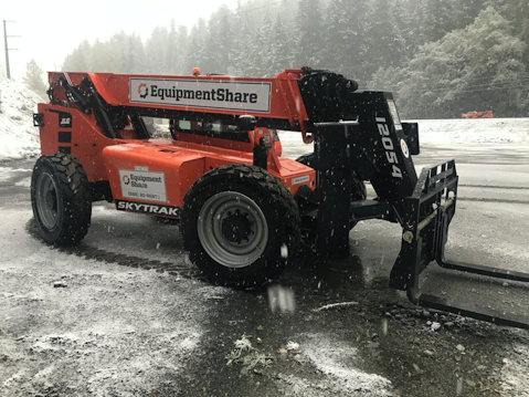 Large orange forklift-style construction vehicle with 'EquipmentShare' branding, parked on a wet and slightly snowy surface, surrounded by a misty forest and power lines in the background.