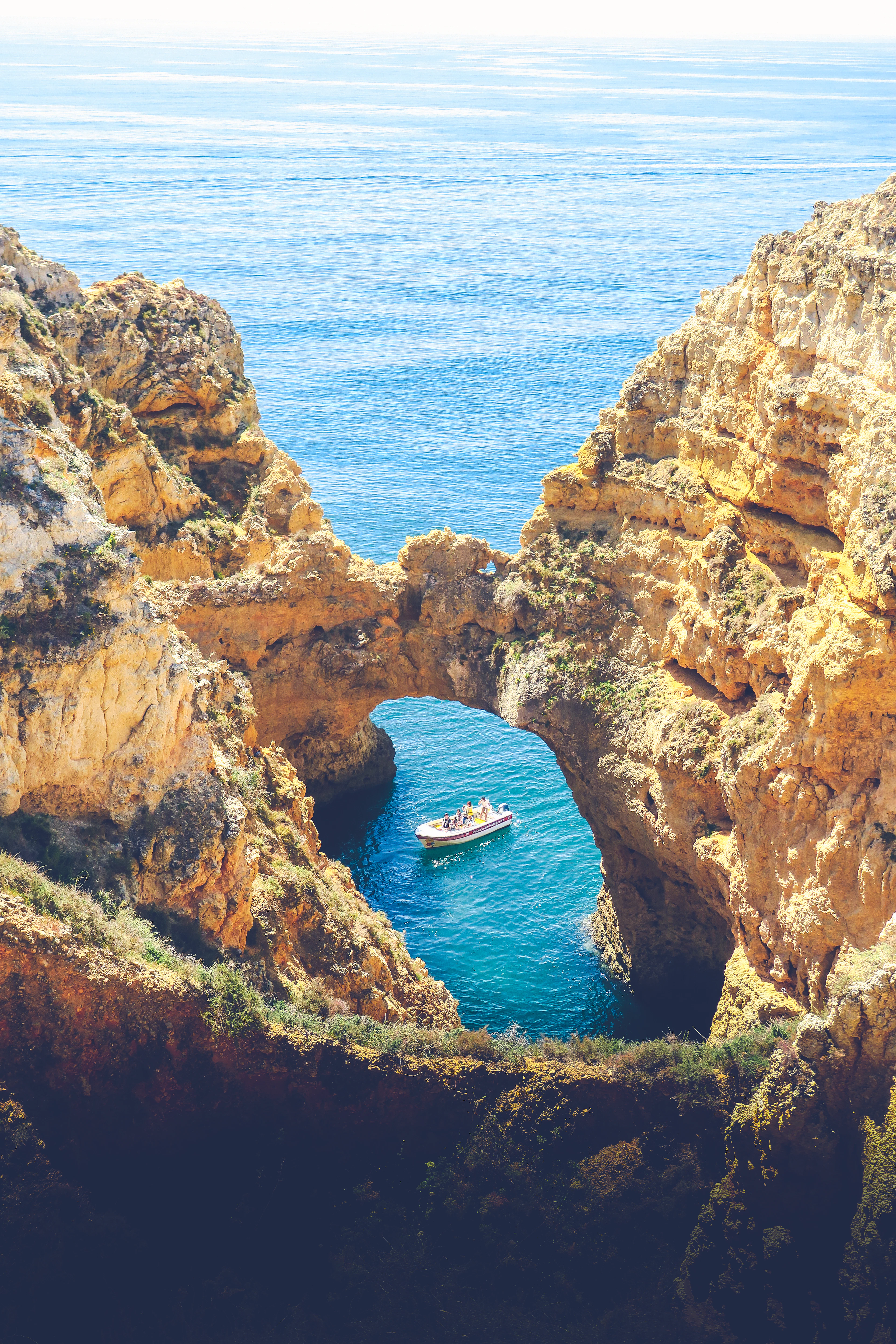 A small boat passes through a natural rock arch in golden sandstone cliffs above blue water.