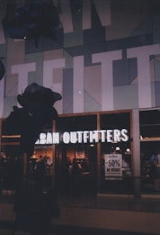 Storefront of Urban Outfitters with illuminated signage and a display window. Inside, clothing and accessories are visible, with a promotional sign offering 50% discount. The surroundings feature geometric wall patterns in muted tones.