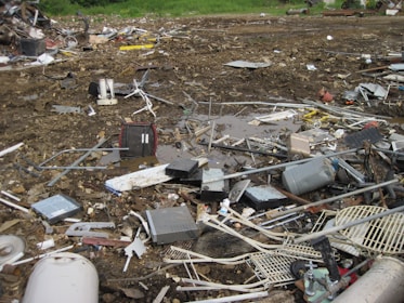 An outdoor scene filled with scattered debris and discarded electronic waste. The ground is covered with broken metal pieces, wires, and various components, creating a cluttered and chaotic atmosphere. In the background, an area with green vegetation is visible.