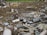 An outdoor scene filled with scattered debris and discarded electronic waste. The ground is covered with broken metal pieces, wires, and various components, creating a cluttered and chaotic atmosphere. In the background, an area with green vegetation is visible.