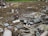 An outdoor scene filled with scattered debris and discarded electronic waste. The ground is covered with broken metal pieces, wires, and various components, creating a cluttered and chaotic atmosphere. In the background, an area with green vegetation is visible.