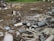 An outdoor scene filled with scattered debris and discarded electronic waste. The ground is covered with broken metal pieces, wires, and various components, creating a cluttered and chaotic atmosphere. In the background, an area with green vegetation is visible.